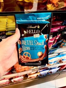 A tall rectangular gradient pink and brown plastic Pouch that has Lindt written in bold gold writing, Hello Pretzel Snack Bites written in bold white writing, and a picture of some medium spherical brown chocolate balls on it, on a white background.