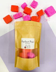 A large rectangular brown paper bag with a white label that has Tales Of Toria Soaps Perfect Pair Bath Rocks written in bold black writing and some pictures of some small rectangular bright pink and orange bath bombs on it, on a white background.