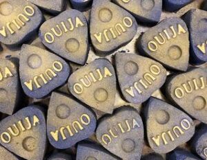 A small dark purple triangular shaped bath bomb that has Ouija written in bold white writing on it, on a white background.