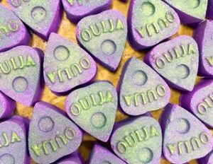 A small light purple triangular shaped bath bomb that has Ouija written in bold white writing on it, on a white background.