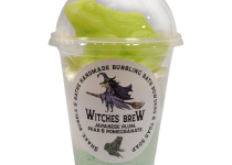 A tall cylindrical clear plastic cup full of some bright green powder with a clear plastic dome lid and a white label that has The Yorkshire Soap Company Witches Brew Shake, Bubble & Bathe written in bold black writing and a picture of a woman wearing a witches outfit on it, on a white background.