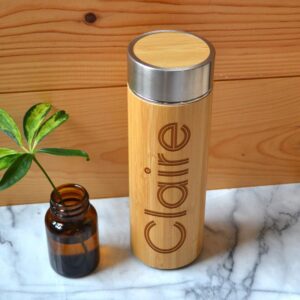 A large cylindrical light wooden bamboo tea flask with a silver metal lid that has Claire engraved on it, on a white background.