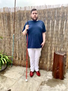 A tall bearded man wearing a dark navy crew neck t-shirt, a pair of bright white chino trousers and a pair of shiny red shoes holding a bright red cane stood against a dark wooden fence.
