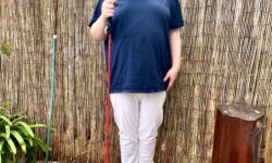 A tall bearded man wearing a dark navy crew neck t-shirt, a pair of bright white chino trousers and a pair of shiny red shoes holding a bright red cane stood against a dark wooden fence.