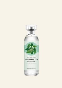 A tall cylindrical clear glass bottle full of some clear liquid with a metallic silver lid and a bright green label that has The Body Shop Fuji Green Tea™ Eau De Cologne written in bold white writing and some pictures of some tea leaves on it, on a white background.