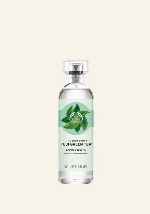 A tall cylindrical clear glass bottle full of some clear liquid with a metallic silver lid and a bright green label that has The Body Shop Fuji Green Tea™ Eau De Cologne written in bold white writing and some pictures of some tea leaves on it, on a white background.