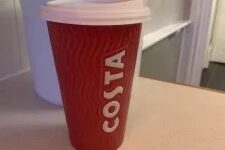 A tall cylindrical red ceramic cup with a white rubber lid that has costa engraved in bold white writing on it, on a white background.