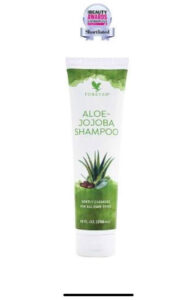 A tall cylindrical White Plastic Bottle with a white plastic lid and a white label that has Forever Living Aloe Jojoba Shampoo written in bright green writing and some pictures of some large dark green aloe vera leaves on it, on a white background.