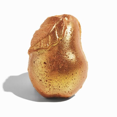 And A Partridge In A Golden Pear Tree | Lush Golden Pear Bath Bomb & Golden Pear Body Spray A large golden orange pear shaped block of solid Body Scrub with a dark brown stem and a cut in the middle of it that’s filled with some bright white cocoa butter, on a white background.