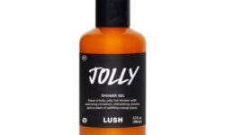 A tall cylindrical clear plastic bottle full of some dark orange liquid with a white label that has Lush Jolly Shower Gel written in bold black writing on it, on a white background.