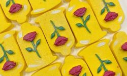 A large bell shaped bright yellow bath bomb that has a bright red rose and a dark green leaf engraved on it, on a white background.