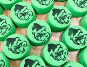 A large circular shaped light green bath bomb that has a pencil drawing of the grinch wearing a black and white Santa’s hat engraved on it, on a white background.