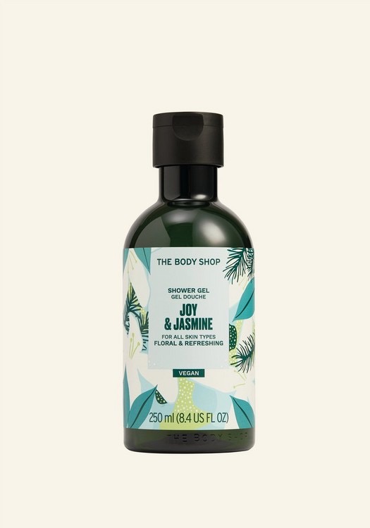 A tall cylindrical clear plastic bottle full of some dark green liquid with a black plastic Lid and a white label that has The Body Shop written in bold black writing, Joy & Jasmine Shower Gel written in bold dark green writing, and some pictures of some large bright green Jasmine leaves on it, on a white background.