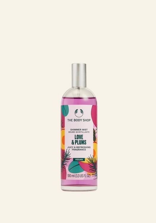 A tall cylindrical clear glass bottle full of some dark pink liquid with a silver metallic lid and a white label that has The Body Shop written in bold black writing, Love & Plums Shimmer Mist written in bold dark green writing, and some pictures of some large dark red plums on it, on a white background.