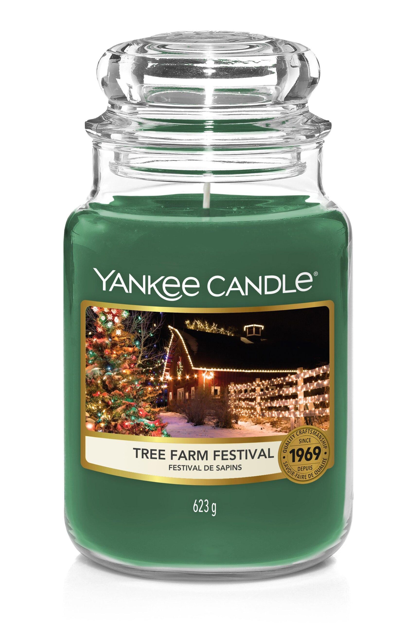 A tall cylindrical glass jar full of some dark green wax with a label that has Yankee Candle written in white writing, Tree Farm Festival written in black writing, and a picture of some bright green Christmas Trees covered in some bright white twinkling lights on it, on a white background.