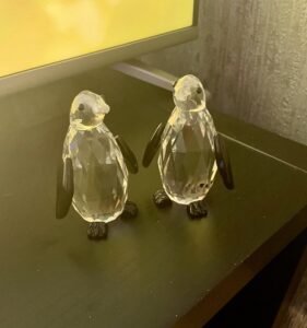Two small black and white crystal penguins in front of a large rectangular black flatscreen Tv, on a black wooden cabinet.