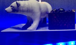 A large rectangular white plate that has a large bright white Polar Bear Tealight holder that’s stood on all four paws next to a small cylindrical glass bowl that’s full of some dark purple and gold wrapped sweets on it and is surrounded by some bright blue strip lights, on a black wooden cabinet.
