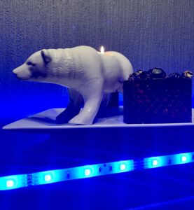 A large rectangular white plate that has a large bright white Polar Bear Tealight holder that’s stood on all four paws next to a small cylindrical glass bowl that’s full of some dark purple and gold wrapped sweets on it and is surrounded by some bright blue strip lights, on a black wooden cabinet.