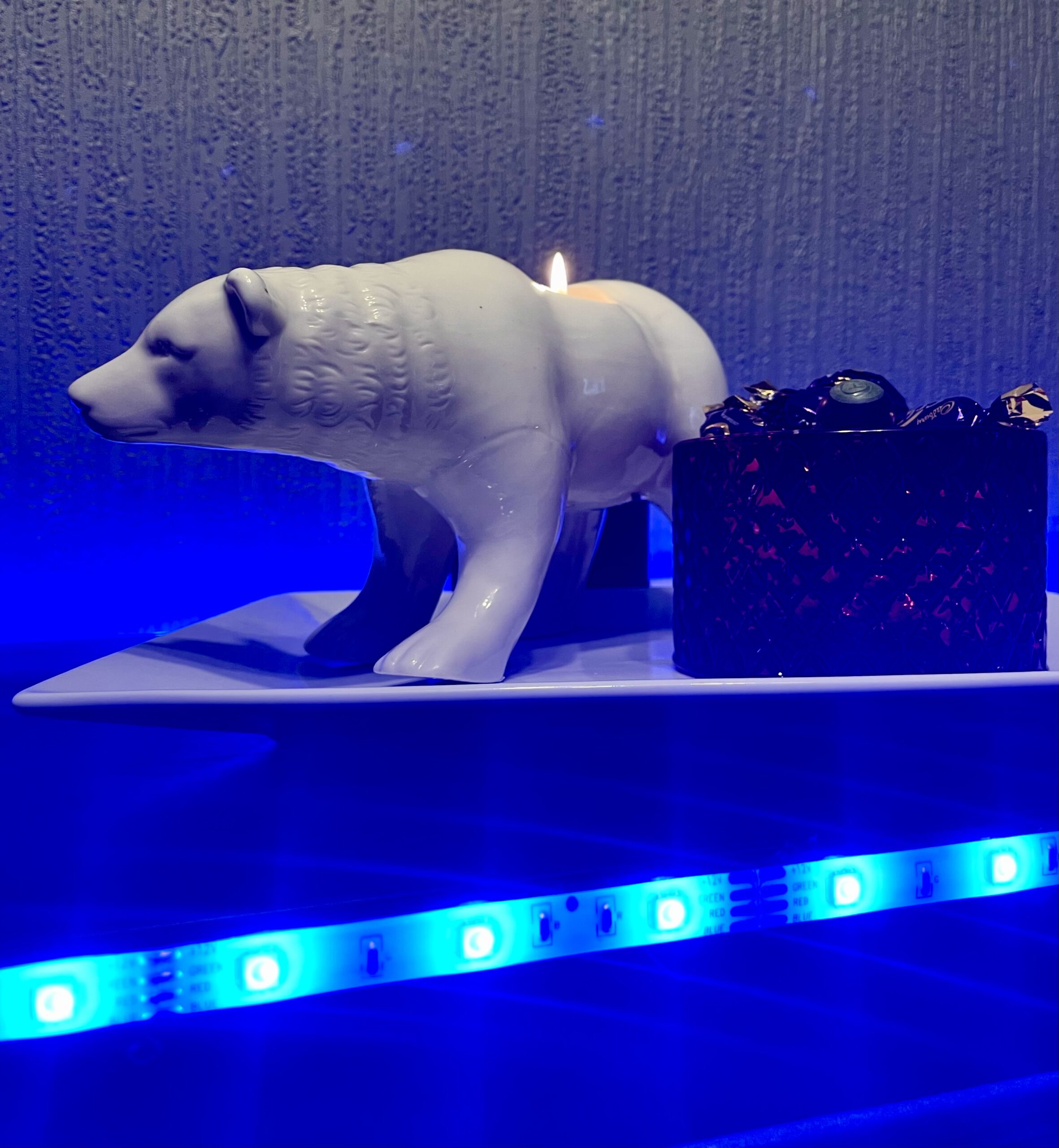 Luke Loves Christmas 2021 | How I've Decorated My Room This Year A large rectangular white plate that has a large bright white Polar Bear Tealight holder that’s stood on all four paws next to a small cylindrical glass bowl that’s full of some dark purple and gold wrapped sweets on it and is surrounded by some bright blue strip lights, on a black wooden cabinet.