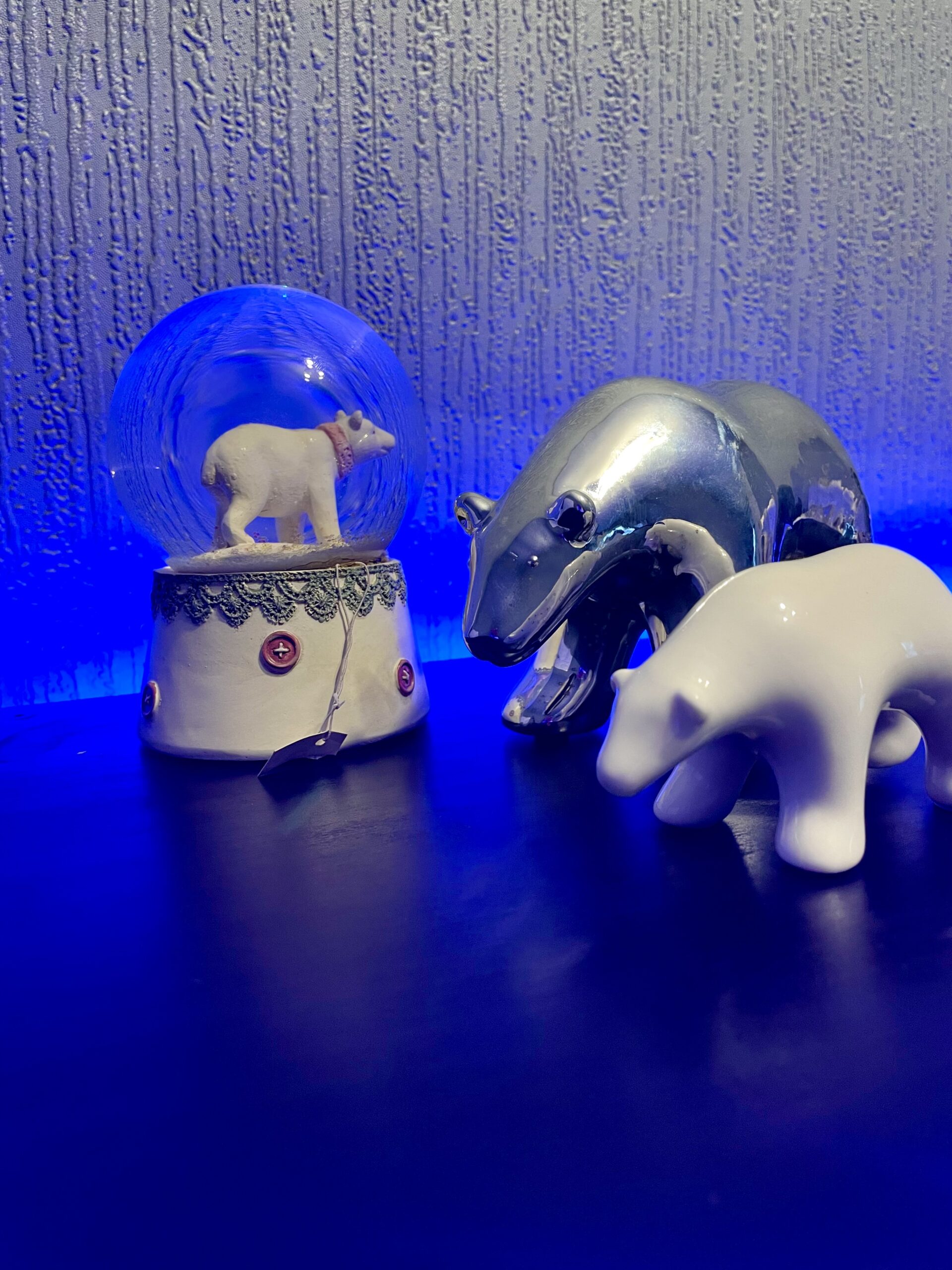 A large bright white spherical snow globe that has a polar bear stood on all four paws inside it next to a medium silver Polar Bear that’s stood on all four paws and a small white polar bear that’s stood on all four paws, on a black wooden cabinet.