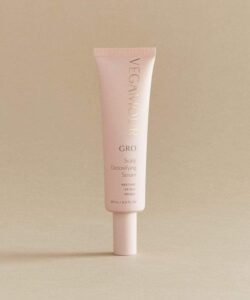 A tall thin conical bright pink plastic tube with a bright pink lid that has Vegamour Grow Scalp Detoxifying Serum written in bold gold writing on it, on a white background.