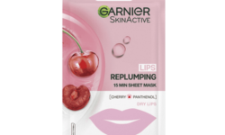 A long and thin gradient pink and white plastic pouch that has Garnier Moisture Bomb Cherry Lip Tissue Mask written in bold black writing and some pictures of some bright red cherries and some bright red lips on it, on a white background.