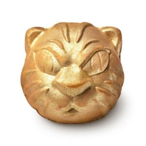 A large pale orange and black striped spherical cat’s head shaped bath bomb that has a tiger’s face engraved on it and some gold lustre on it, on a white background.