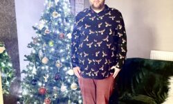 A tall bearded man wearing a black long sleeve shirt that has a white and red moose print all over it, a dark red V Neck t-shirt, a pair of dark red chinos and a pair of shiny red shoes stood in front of a large dark green Christmas Tree that has some twinkling bright white lights all over it.