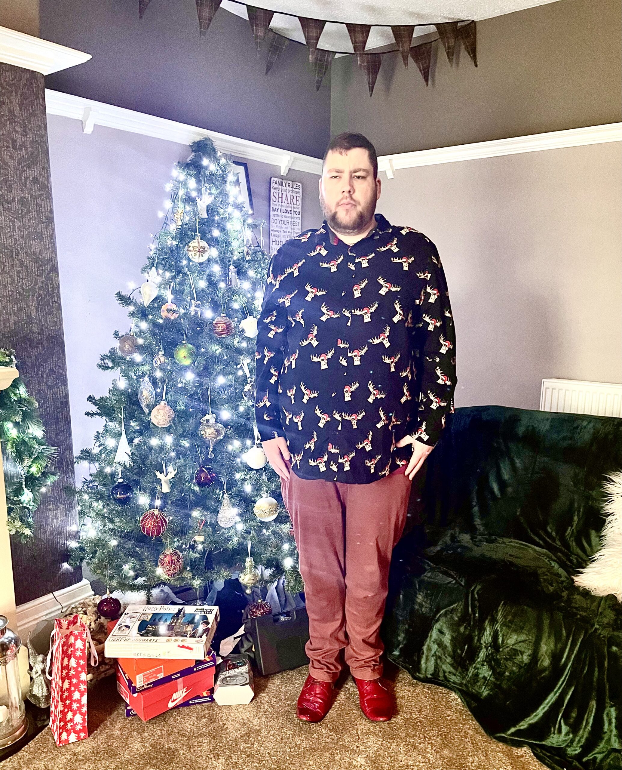 Outfit Of The Day | Rudolph The Red Nosed Reindeer A tall bearded man wearing a black long sleeve shirt that has a white and red moose print all over it, a dark red V Neck t-shirt, a pair of dark red chinos and a pair of shiny red shoes stood in front of a large dark green Christmas Tree that has some twinkling bright white lights all over it.