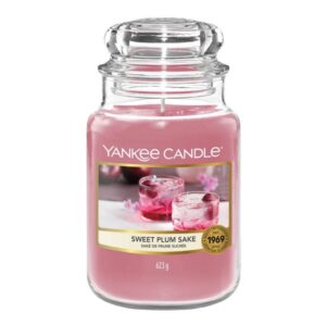A tall cylindrical glass jar full of some bright pink wax with a label that has Yankee Candle written in black writing, Sweet Plum Sake written in black writing, and a picture of a tall glass full of some dark brown liquid and some large dark red plums on it, on a white background.