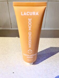 A tall conical bright orange plastic tube with a white plastic lid that has Aldi Weekend Mode Exfoliating Mask written in bold white writing on it, on a white background