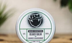 A short cylindrical silver metal tin with a white label that has Beard Monsters Liquorice & Blackcurrant Beard Balm written in bold black writing and a picture of a man with a black beard on it, on a white background.