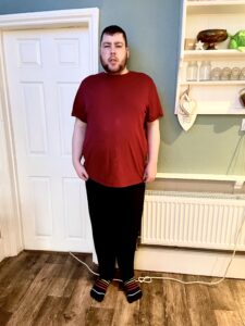 A tall bearded man wearing a dark red crew neck t-shirt, a pair of black bamboo lounge bottoms, and a pair of black and multi-coloured striped socks stood against a light green wall.