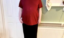 A tall bearded man wearing a dark red crew neck t-shirt, a pair of black bamboo lounge bottoms, and a pair of black and multi-coloured striped socks stood against a light green wall.