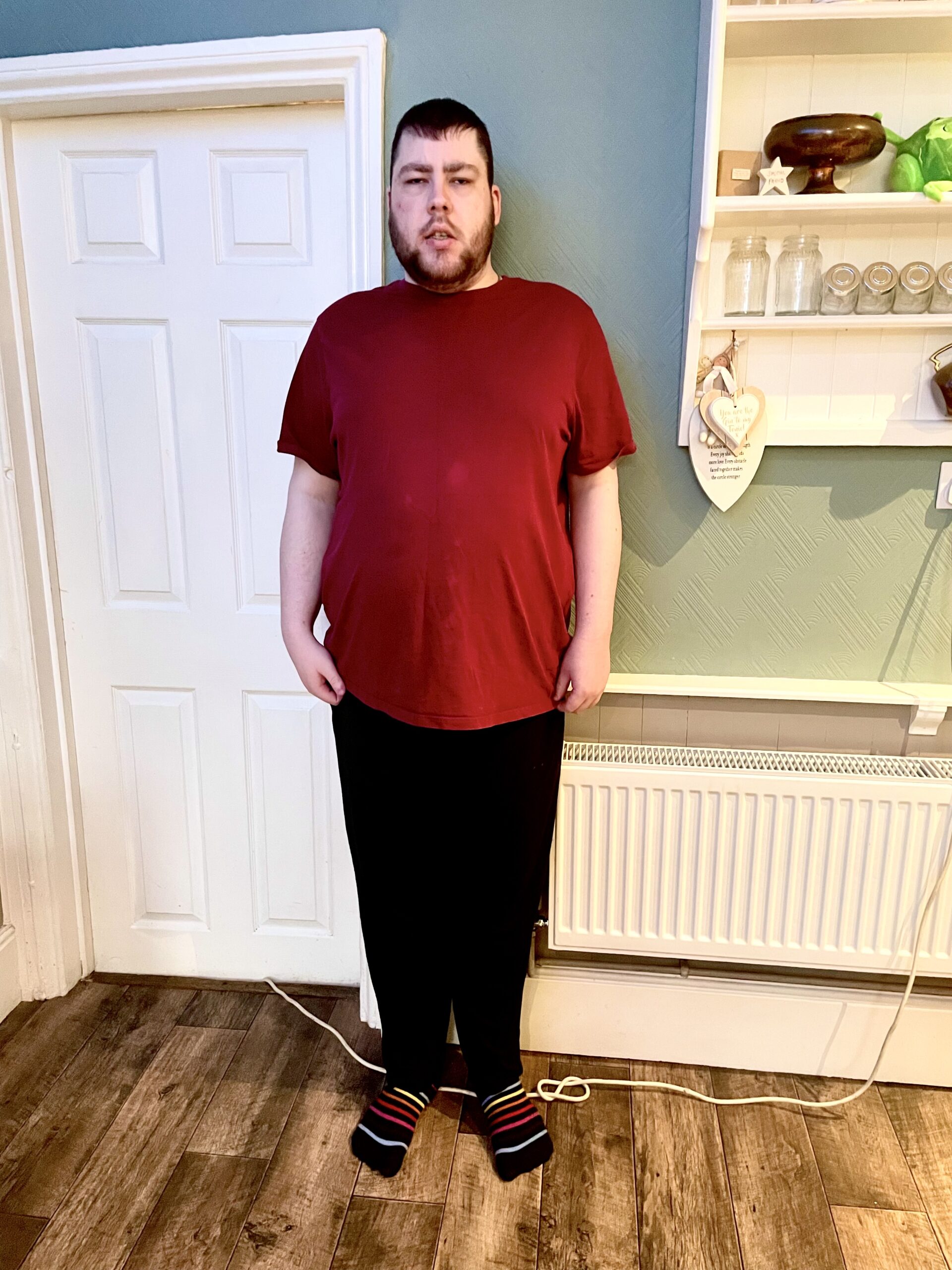 I Look Like One Lazy Panda When I'm Lounging Around The House | SockShop Men's Lazy Panda Bamboo Loungewear Selection Classic Bottoms A tall bearded man wearing a dark red crew neck t-shirt, a pair of black bamboo lounge bottoms, and a pair of black and multi-coloured striped socks stood against a light green wall.