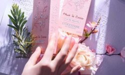 A tall rectangular white cardboard box that has L'Occitane Fleurs De Crisier written in bold black cursive writing and some braille dots on it covered by a large light pink hand, on a white background.