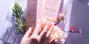 A tall rectangular white cardboard box that has L'Occitane Fleurs De Crisier written in bold black cursive writing and some braille dots on it covered by a large light pink hand, on a white background.