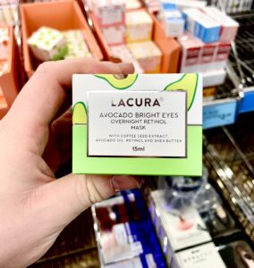 A hand holding a tall rectangular white cardboard box that has Aldi Lacura Avocado Bright Eyes Overnight Retinol Sleeping Mask written in bold black writing, a picture of a dark green stripe, and some pictures of some dark green avocados on it, on a white background.