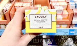 A hand holding a tall rectangular white cardboard box that has Aldi Lacura Banana Soufflé Face Cream written in bold black writing, a picture of a bright yellow stripe, and some pictures of some bright yellow bananas on it, on a white background.