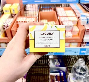A hand holding a tall rectangular white cardboard box that has Aldi Lacura Banana Soufflé Face Cream written in bold black writing, a picture of a bright yellow stripe, and some pictures of some bright yellow bananas on it, on a white background.