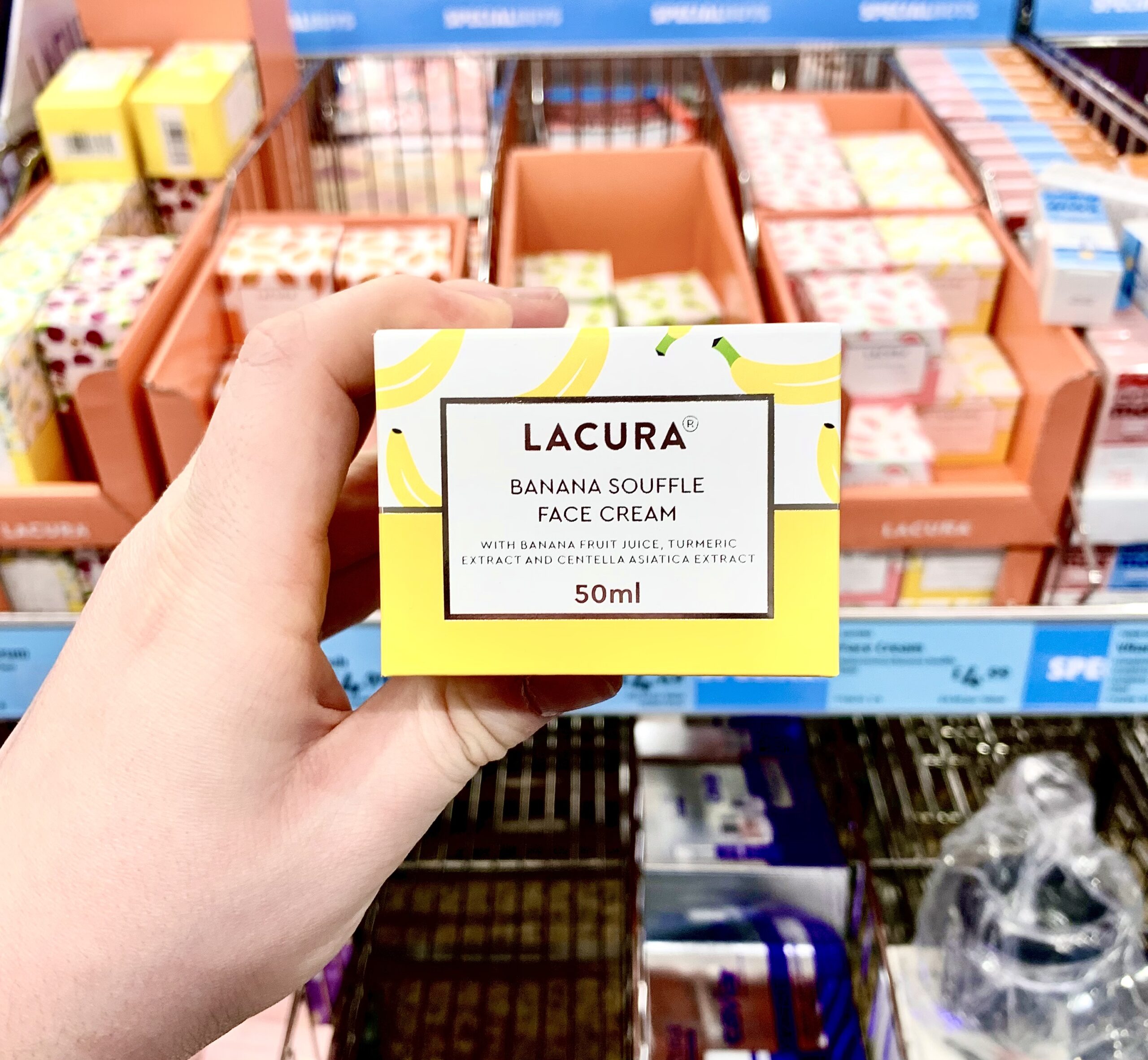 A hand holding a tall rectangular white cardboard box that has Aldi Lacura Banana Soufflé Face Cream written in bold black writing, a picture of a bright yellow stripe, and some pictures of some bright yellow bananas on it, on a white background.