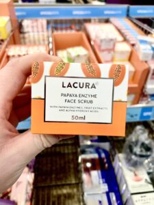 A hand holding a short rectangular white cardboard box that has Aldi Lacura Papaya Enzyme Face Scrub written in bold black writing, a picture of a dark orange stripe, and some pictures of some dark orange Papayas on it, on a white background.