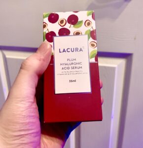 A hand holding a tall rectangular white cardboard box that has Aldi Lacura Plum Hyaluronic Acid Serum written in bold black writing, a picture of a dark Plum stripe, and some pictures of some dark red Plums on it, on a white background.