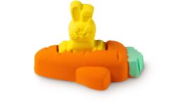 A large bright orange carrot shaped bath bomb that has a small bright yellow rabbit shaped bath bomb coming out of the top of it and some bright green leaves coming out of the side of it, on a white background.