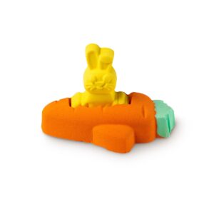 A large bright orange carrot shaped bath bomb that has a small bright yellow rabbit shaped bath bomb coming out of the top of it and some bright green leaves coming out of the side of it, on a white background.