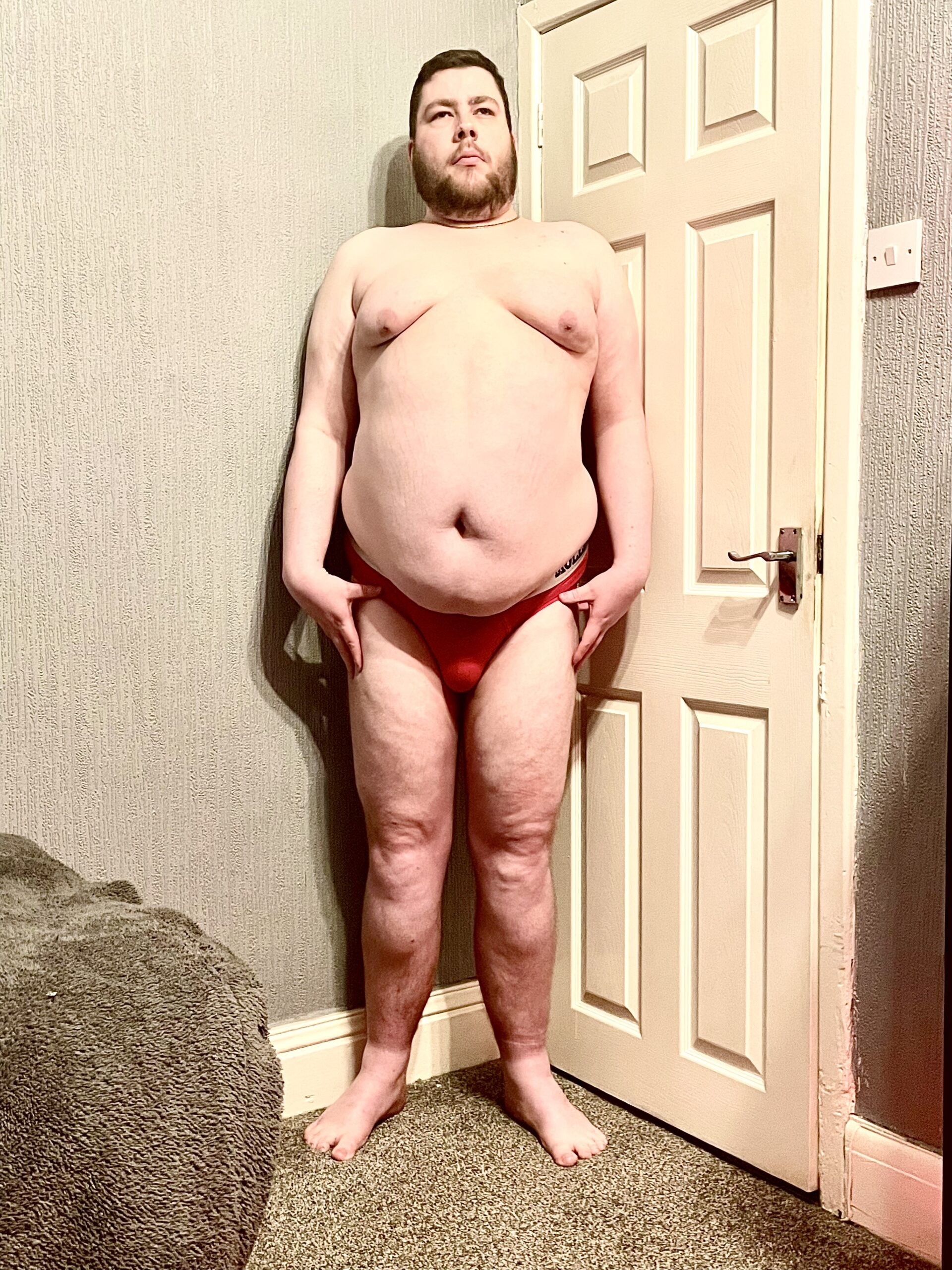 A tall bearded man wearing a pair of bright Red Mesh Briefs with a bright Red Cotton Pouch and a bright White Waistband that has Molly & Tommy written in bold red writing on it stood against a white wooden door.