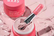 A short cylindrical bright Pink glass jar full of some creamy pink liquid with a bright pink plastic face mask brush coming out of the top of it that has Sand & Sky Australian Pink Clay Porefining Face Mask written in bold white writing on it, on a white background.