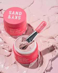 A short cylindrical bright Pink glass jar full of some creamy pink liquid with a bright pink plastic face mask brush coming out of the top of it that has Sand & Sky Australian Pink Clay Porefining Face Mask written in bold white writing on it, on a white background.