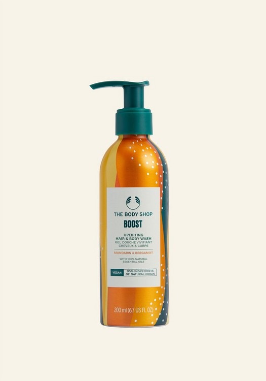 Taking Care Of My Wellbeing Whilst I Have A Shower | The Body Shop Wellness Range A tall cylindrical metallic Orange Aluminium Bottle with a Silver Aluminium lid and a white label that has The Body Shop Boost Uplifting Hair & Body Wash written in dark green writing and some pictures of some bright Orange swirls on it, on a white background.