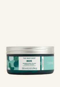 A short cylindrical clear glass jar full of some dark green liquid with a silver Aluminium Lid and a white label that has The Body Shop Breathe Whisked Body Polish written in dark green writing and some pictures of some light green swirls on it, on a white background.